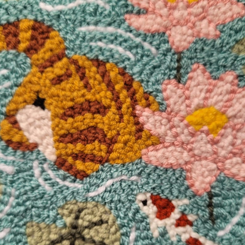 May include: A punch needle textile art piece featuring a textured teal background with white water ripples. The design includes a golden-brown tiger, two light pink lotus flowers with yellow centers, a small white and red koi fish, and green lily pads.