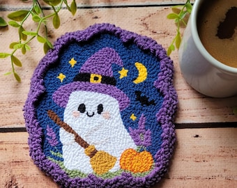 May include: A punch needle coaster featuring a white ghost wearing a purple witch hat, holding a brown broomstick, and standing next to an orange pumpkin. The background is a dark blue night sky with yellow stars, a crescent moon, and a black bat.