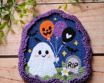May include: A punch needle wall hanging featuring a white ghost holding three balloons in orange, purple, and black. The scene includes a gray tombstone with the text RIP, small yellow stars, and black bats against a dark blue background, all framed by a textured purple border.
