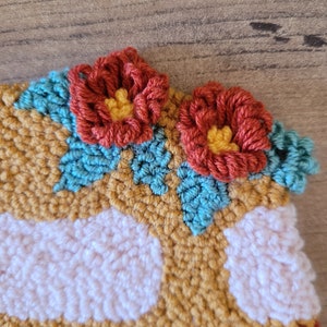 Op de afbeelding: Een punch needle textielstuk met een mosterdgele achtergrond, witte cirkelvormige patronen en twee driedimensionale roestkleurige bloemen met gele harten, geaccentueerd door kleine blauwgroene blaadjes die in het textuurrijke oppervlak zijn gestikt.