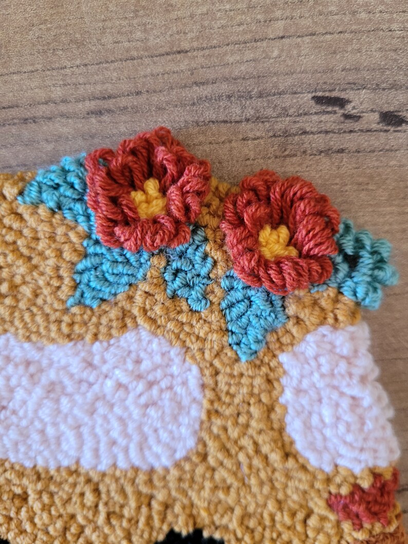 Op de afbeelding: Een punch needle textielstuk met een mosterdgele achtergrond, witte cirkelvormige patronen en twee driedimensionale roestkleurige bloemen met gele harten, geaccentueerd door kleine blauwgroene blaadjes die in het textuurrijke oppervlak zijn gestikt.