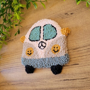 Op de afbeelding: Een punch needle onderzetter in de vorm van een busje in de kleuren cr&egrave;me, lichtgrijs en donkergrijs. Het busje heeft twee blauwgroene ramen, een vredesteken, twee gele smiley-iconen en twee zwarte wielen met kleine oranje zijspiegels.