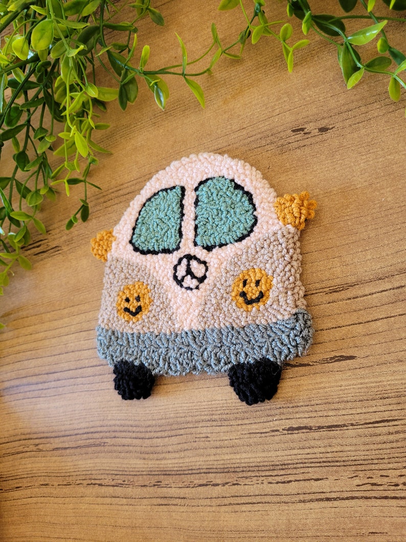 Op de afbeelding: Een punch needle onderzetter in de vorm van een busje in de kleuren cr&egrave;me, lichtgrijs en donkergrijs. Het busje heeft twee blauwgroene ramen, een vredesteken, twee gele smiley-iconen en twee zwarte wielen met kleine oranje zijspiegels.