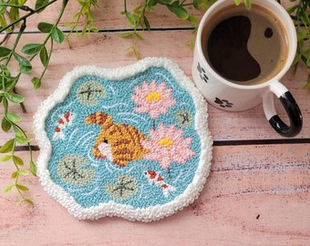 May include: A punch needle coaster with a light blue background featuring a brown tabby cat, two pink lotus flowers, green lily pads, and two small koi fish. The coaster has a white textured border and sits next to a white ceramic mug.