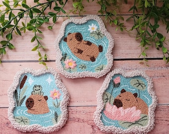 May include: Three punch needle coasters featuring capybaras in a pond. One shows a capybara swimming, another a capybara with a frog, and the third a capybara resting on a pink lotus flower. Each coaster has a light blue background and cream border.