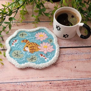 May include: A punch needle coaster featuring a light blue pond with a brown tabby cat rear, two pink lotus flowers, green lily pads, and two small koi fish. The coaster has a white textured border and is paired with a white mug with black paw prints.