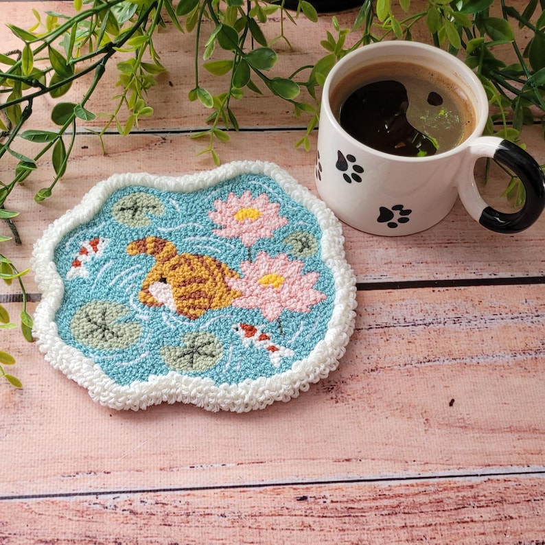 May include: A punch needle coaster featuring a light blue pond with a brown tabby cat rear, two pink lotus flowers, green lily pads, and two small koi fish. The coaster has a white textured border and is paired with a white mug with black paw prints.
