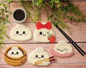 May include: Five tufted coasters featuring cute, embroidered faces on dumpling-shaped designs. One has a red bow and strawberry, one is in a brown bamboo steamer, one holds chopsticks, and one has pink accents. A small white cup of dark liquid is included.