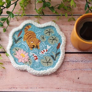 May include: A punch needle coaster with a light blue background featuring a ginger cat swimming, two white and orange koi fish, a pink water lily, and green lily pads. The coaster has a thick, white, textured border and an irregular shape.