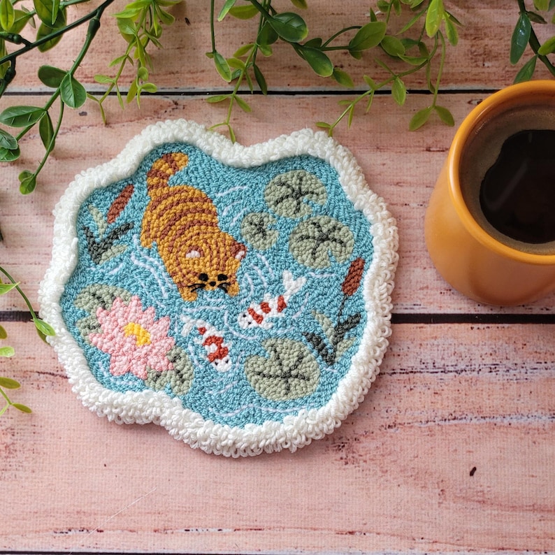 May include: A punch needle coaster with a light blue background featuring a ginger cat swimming, two white and orange koi fish, a pink water lily, and green lily pads. The coaster has a thick, white, textured border and an irregular shape.