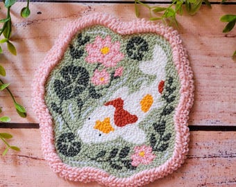 May include: A punch needle coaster featuring a white koi fish with orange and red patches swimming in a sage green pond. The design includes dark green lily pads and pink lotus flowers, all bordered by a thick, textured light pink yarn edge.