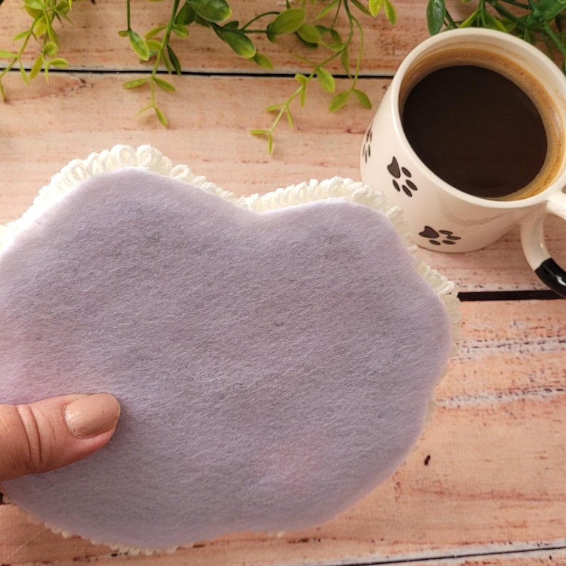 May include: A heart-shaped coaster featuring a smooth, light purple felt top surface and a textured, white crocheted border. The coaster is positioned next to a white ceramic mug decorated with black paw prints and filled with dark coffee.