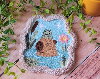 May include: A punch needle coaster featuring a brown capybara in light blue water with a small green frog on its head. The design includes a pink flower, green cattails, and white star accents, all bordered by a textured, beige looped yarn edge.