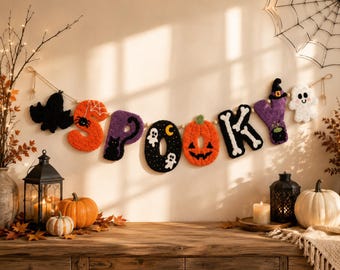 May include: A decorative wall garland spelling the word SPOOKY, featuring plush letters in orange, purple, and black. The letters are adorned with embroidered Halloween motifs including a spider, a black cat, ghosts, a crescent moon, a pumpkin face, and bones.