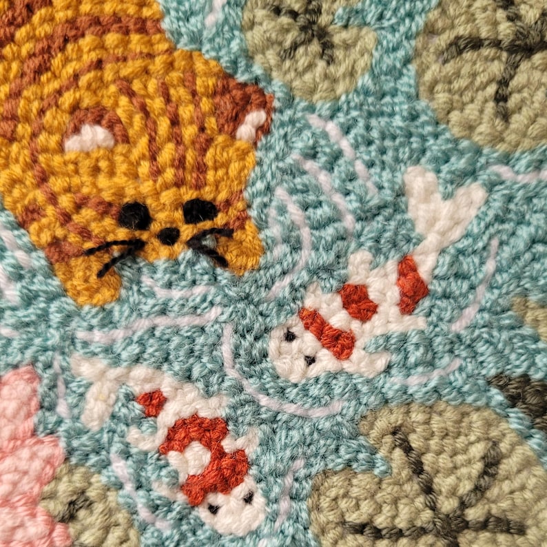 May include: A punch needle textile art piece featuring a golden orange tabby cat face looking down at two white and orange koi fish swimming in light blue water surrounded by green lily pads and a pink flower in the corner.
