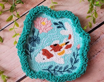 May include: A punch needle coaster featuring a white koi fish with orange and yellow spots swimming in light blue water. The design includes a pink lotus flower and dark green aquatic plants, finished with a textured teal crocheted border.