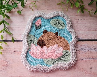 May include: A punch needle textile art piece featuring a brown capybara resting inside a large pink lotus flower. The background is light blue with green lily pads and a pink lotus bud, all surrounded by a thick, cream-colored looped yarn border.