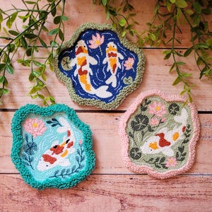 May include: Three irregular, hand-tufted coasters featuring koi fish and floral motifs. One coaster has a dark blue background with two koi, one has a light blue background with one koi, and one has a sage green background with one koi.