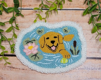 May include: A punch needle coaster featuring a golden dog swimming in a blue pond with a pink water lily, green lily pads, and a cattail. The dog has its mouth open and is reaching for a yellow ball. The piece has a white crocheted border.
