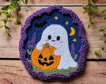 May include: A punch needle coaster featuring a white ghost holding an orange jack-o'-lantern against a dark blue background. The design includes a yellow crescent moon, small yellow stars, a black bat, and purple accents, all framed by a purple border.