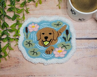 May include: A rectangular punch needle coaster featuring a brown dog swimming in blue water with a yellow ball in its mouth. The scene includes pink water lilies, green lily pads, and brown cattails, all bordered by a white crocheted scalloped edge.