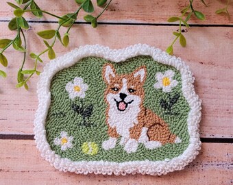 May include: A rectangular punch needle coaster featuring a brown and white Corgi dog sitting on a green background with white flowers and a yellow tennis ball. The coaster has a thick, textured white border with a scalloped edge design.