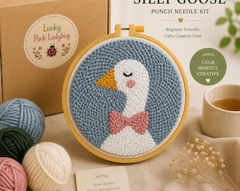 May include: A punch needle kit featuring a brown cardboard box labeled SILLY GOOSE, a wooden embroidery hoop with a white goose design on a blue background, four yarn balls in blue, green, cream, and pink, and a card with the text Slow down.