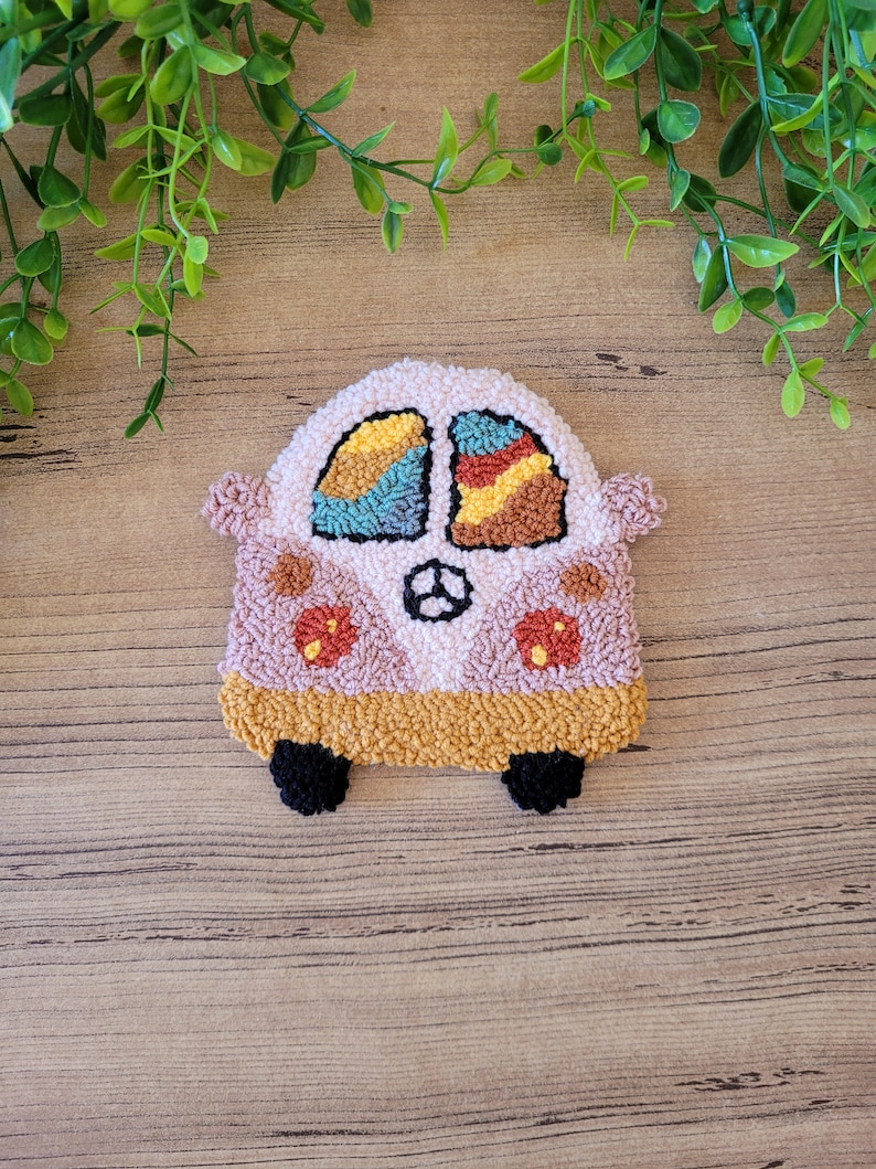 Op de afbeelding: Een punch needle onderzetter in de vorm van een busje in tinten lichtroze, mosterdgeel en zwart. Het busje heeft twee ramen met veelkleurige garenpatronen, een centraal stuurwiel-embleem en twee ronde rode en oranje bloemaccenten.