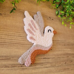 Op de afbeelding: Een punch needle wandhanger in de vorm van een vogel met een cr&egrave;mekleurig lichaam, een bruine snavel en een gesloten oog. De vleugels en staart tonen gelaagde secties van cr&egrave;me, lichtbeige, oudroze en terracottakleurig garen met textuur.