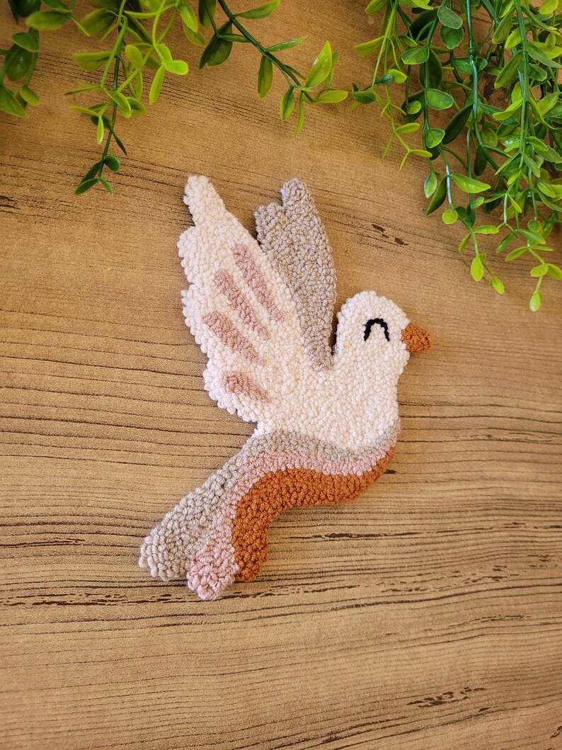 Op de afbeelding: Een punch needle wandhanger in de vorm van een vogel met een cr&egrave;mekleurig lichaam, een bruine snavel en een gesloten oog. De vleugels en staart tonen gelaagde secties van cr&egrave;me, lichtbeige, oudroze en terracottakleurig garen met textuur.