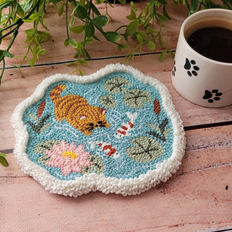 May include: A punch needle coaster featuring a light blue pond scene with a ginger tabby cat, two koi fish, a pink water lily, and green lily pads. The coaster has an irregular shape with a textured white crocheted border edge.