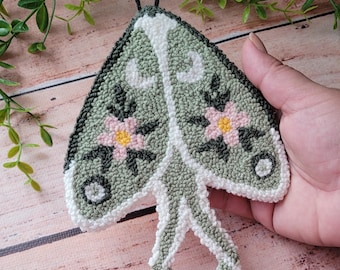 May include: A punch needle textile art piece shaped like a moth with sage green wings, white borders, and two black antennae. The wings feature embroidered pink flowers with yellow centers, dark green leaves, and white crescent moon accents.
