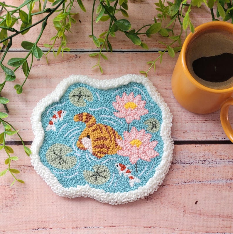 May include: A tufted coaster with a light blue background featuring a brown striped cat, two pink lotus flowers, green lily pads, and two small koi fish. The coaster has an irregular, cloud-like shape with a thick, white, textured border.