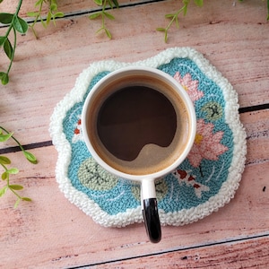 May include: A white ceramic mug filled with black coffee sits on a round, tufted coaster. The coaster features a light blue background with embroidered pink lotus flowers, green lily pads, and small orange and white koi fish, finished with a white border.