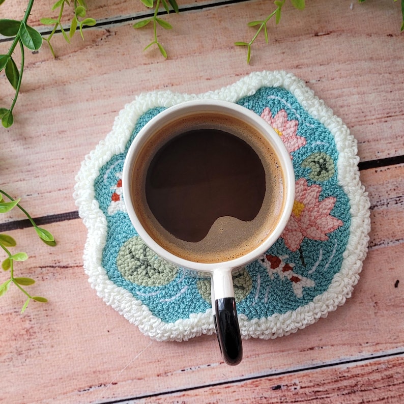 May include: A white ceramic mug filled with black coffee sits on a round, tufted coaster. The coaster features a light blue background with embroidered pink lotus flowers, green lily pads, and small orange and white koi fish, finished with a white border.