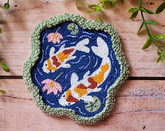 May include: A punch needle coaster featuring two koi fish in white, orange, and brown swimming in a dark blue pond. The design includes pink lotus flowers and green lily pads, all surrounded by a thick, textured sage green crocheted border.