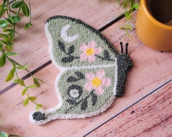 May include: A punch needle butterfly coaster in sage green with a white crescent moon, dark green leafy stems, and two pink flowers with yellow centers. The butterfly shape features a dark gray body and a white border, crafted with textured yarn loops.