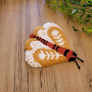 Op de afbeelding: Een punch needle textielkunstwerk in de vorm van een mot met gestructureerde vleugels in mosterdgeel en wit. De mot heeft een centraal lichaam in roestoranje met zwarte horizontale strepen en twee dunne zwarte voelsprieten die uitsteken.