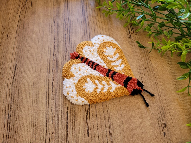Op de afbeelding: Een punch needle textielkunstwerk in de vorm van een mot met gestructureerde vleugels in mosterdgeel en wit. De mot heeft een centraal lichaam in roestoranje met zwarte horizontale strepen en twee dunne zwarte voelsprieten die uitsteken.
