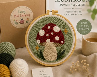 May include: A punch needle kit featuring a mushroom design in a wooden hoop. The kit includes a brown cardboard box labeled Lucky Pink Ladybug, balls of yarn in green, dark green, and cream, and a card with the text Slow down Enjoy the process You've got this. The mushroom design has a red cap with white spots, a cream stem, and green leaves on a green background.