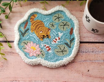 May include: A punch needle coaster with a light blue background featuring an orange tabby cat, two koi fish, a pink water lily, and green lily pads. The coaster has a thick, white, textured border and a wavy, irregular shape.