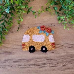 Op de afbeelding: Een punch needle handwerkstuk in de vorm van een camper in mosterdgeel met lichtroze ramen, zwarte wielen en twee rode bloemen met blauwgroene bladeren op het dak. Het getextureerde textielontwerp heeft een dichte constructie van lussen.