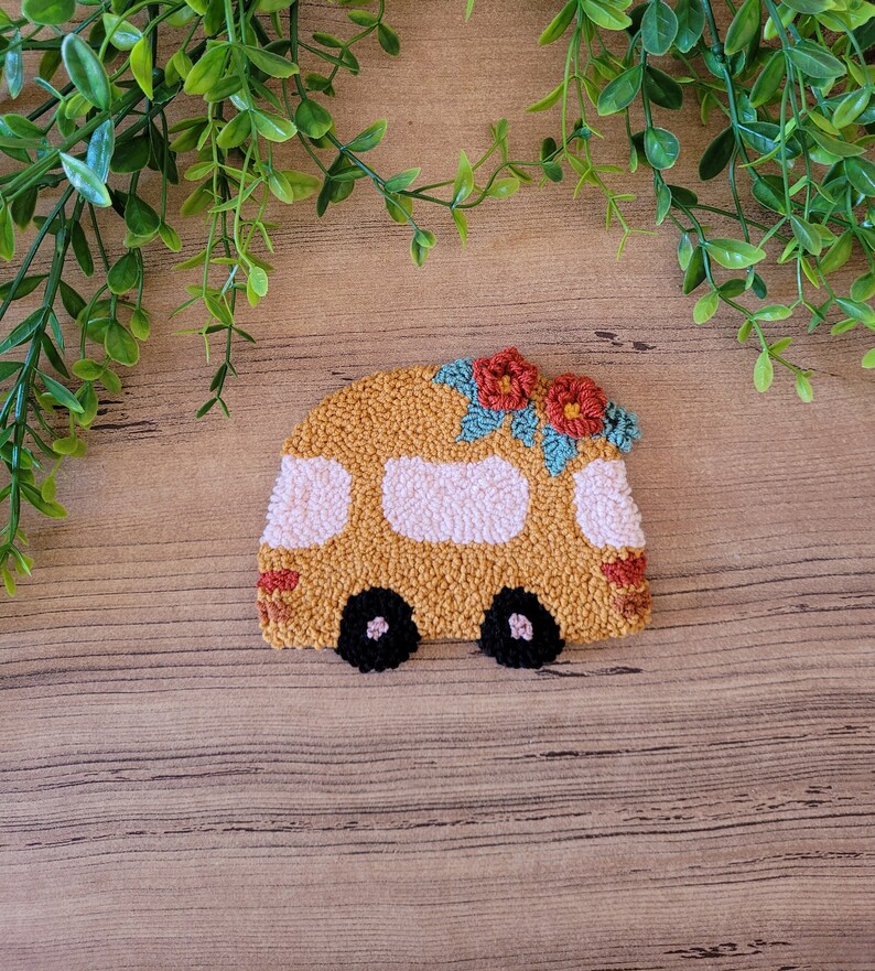 Op de afbeelding: Een punch needle handwerkstuk in de vorm van een camper in mosterdgeel met lichtroze ramen, zwarte wielen en twee rode bloemen met blauwgroene bladeren op het dak. Het getextureerde textielontwerp heeft een dichte constructie van lussen.