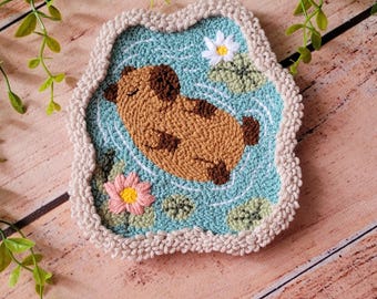 May include: A punch needle coaster featuring a brown capybara floating in a light blue pond with white and pink water lilies and green lily pads. The coaster has a thick, textured beige crocheted border and a soft, looped yarn surface.