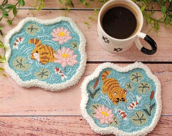 May include: Two tufted coasters with a light blue background featuring a pond scene with pink lotus flowers, green lily pads, small koi fish, and an orange tabby cat. The coasters have a white, textured, wavy border and a soft, looped pile finish.