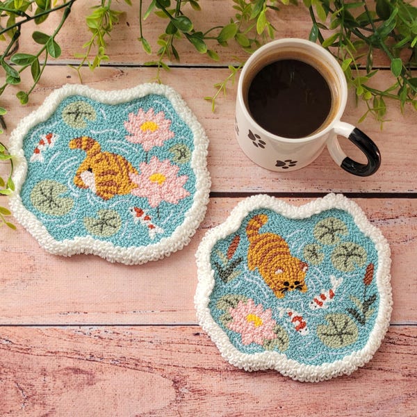 Puede incluir: Dos posavasos de tufting con fondo azul claro que muestran una escena de estanque con flores de loto rosas, nenúfares verdes, pequeños peces koi y un gato atigrado naranja. Los posavasos tienen un borde blanco texturizado y ondulado.