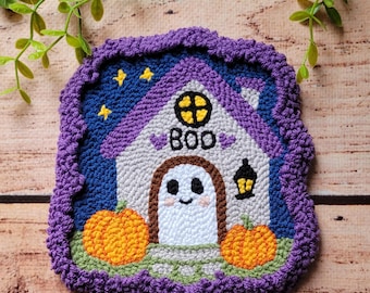 May include: A square punch needle coaster featuring a white house with a purple roof, a ghost in the doorway, and the text "BOO" above it. Two orange pumpkins sit on a green patch, set against a dark blue night sky with yellow stars.