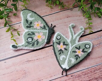 May include: Two punch needle textile art pieces shaped like butterflies and moths in sage green, featuring white crescent moon accents, dark green leaf embroidery, and small pink flowers with yellow centers, crafted with a textured yarn finish.