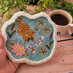 May include: A circular punch needle coaster featuring a blue pond scene with a ginger tabby cat, two white and orange koi fish, a pink water lily, and green lily pads. The coaster has a thick, textured white yarn border and a soft, looped finish.