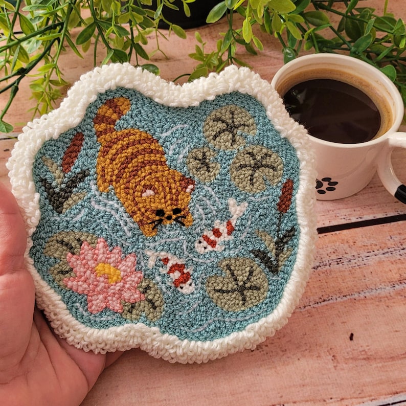 May include: A circular punch needle coaster featuring a blue pond scene with a ginger tabby cat, two white and orange koi fish, a pink water lily, and green lily pads. The coaster has a thick, textured white yarn border and a soft, looped finish.
